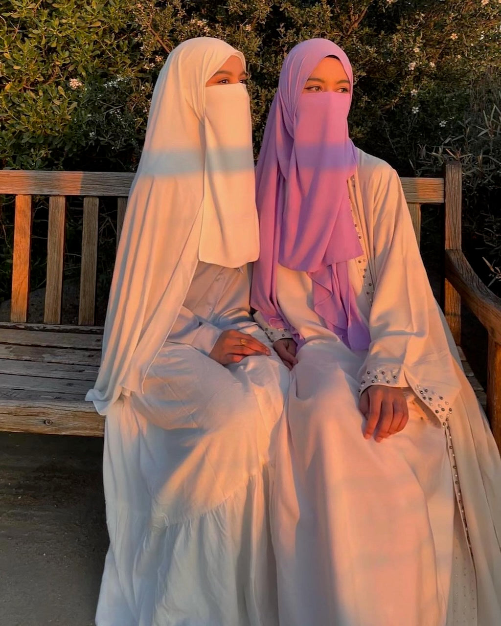 Two women wearing modest white and lavender abayas and face veils sit side by side on a wooden bench outdoors at sunset, with warm golden light casting shadows across their garments.