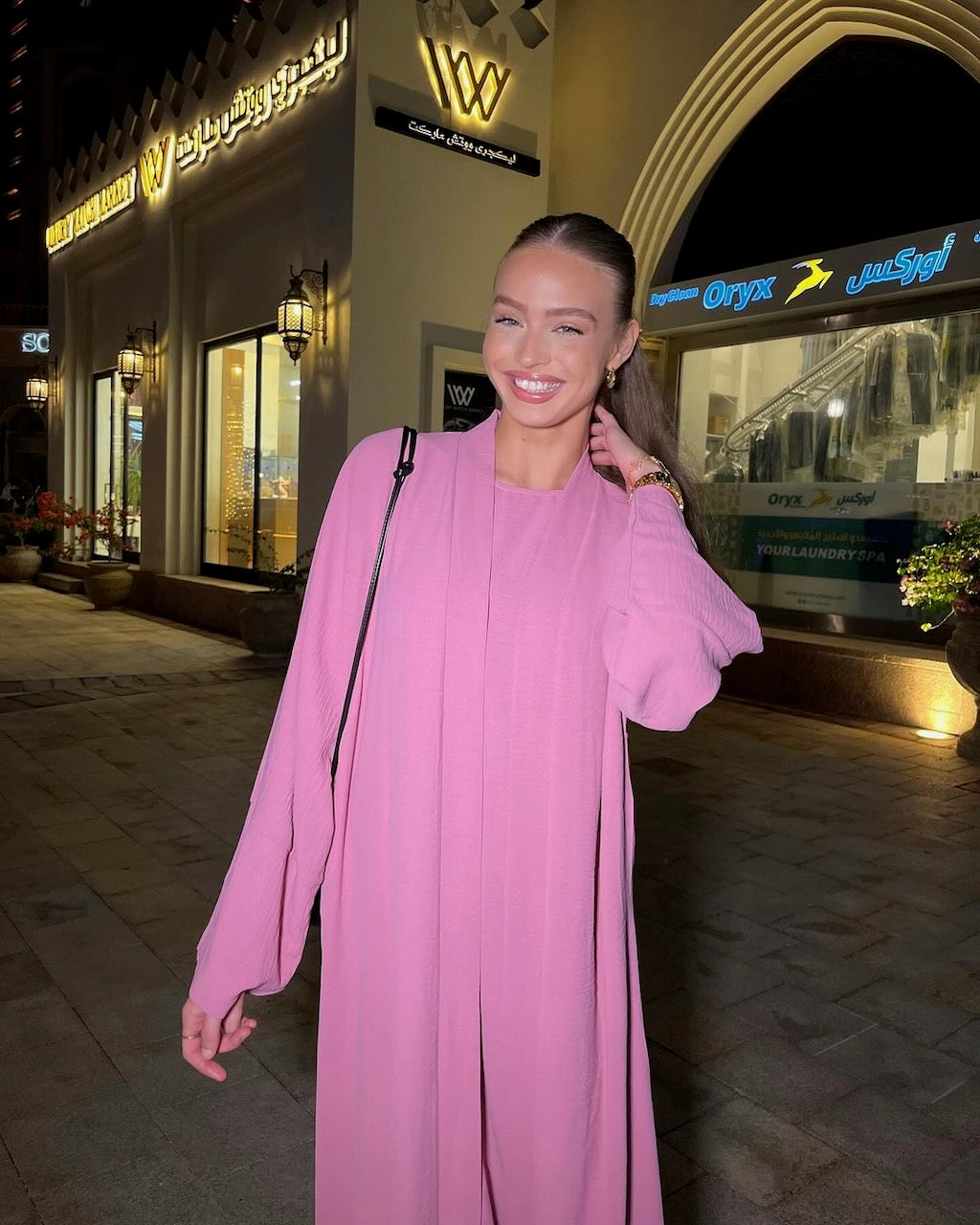 A woman wearing a long, flowing pink abaya with voluminous sleeves, standing outdoors in front of a lit-up shopping center at night.