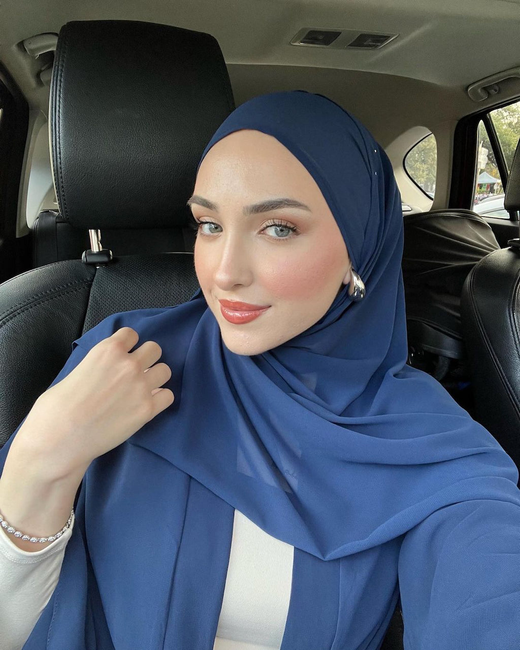 A woman wearing a solid navy blue chiffon hijab and navy aisha chiffon abaya sits in a black leather car seat, posing for a selfie.