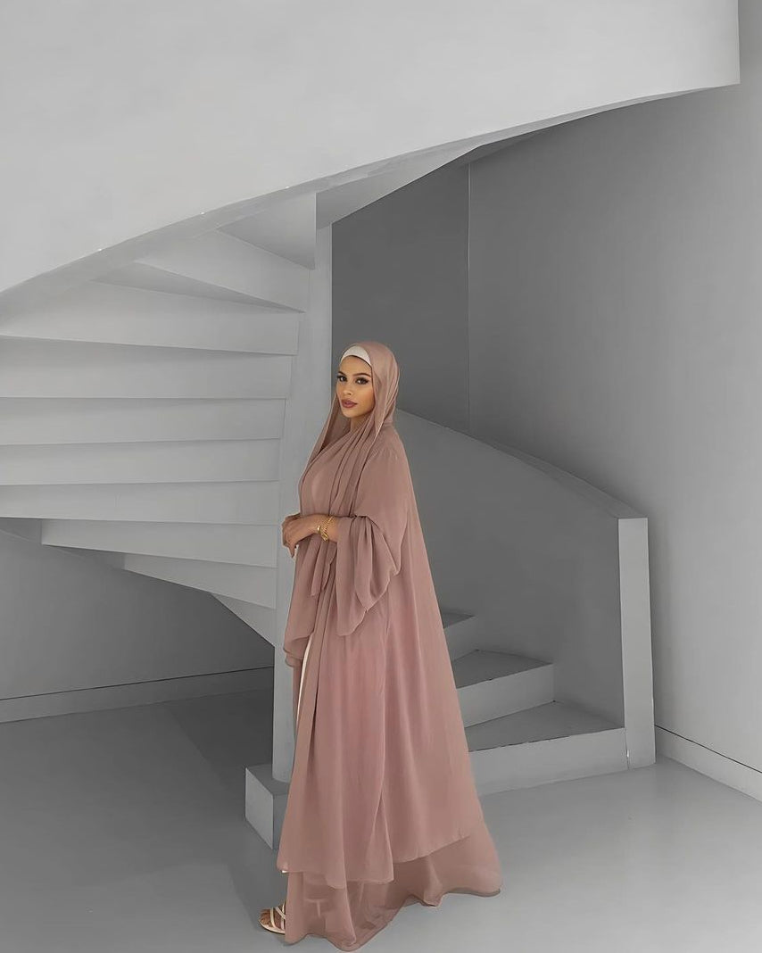 A woman wearing a flowing dusty rose aisha nude abaya and matching hijab stands on a minimalist white spiral staircase.