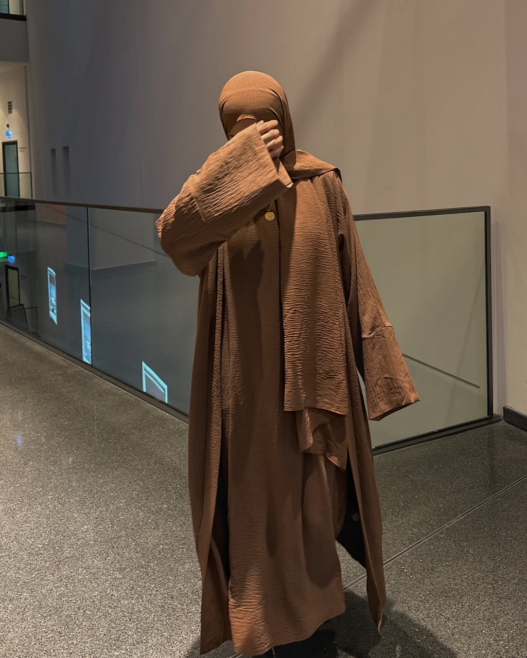 A person wearing a long, flowing brown abaya and matching head covering stands indoors near a glass railing, with one hand covering part of the face.