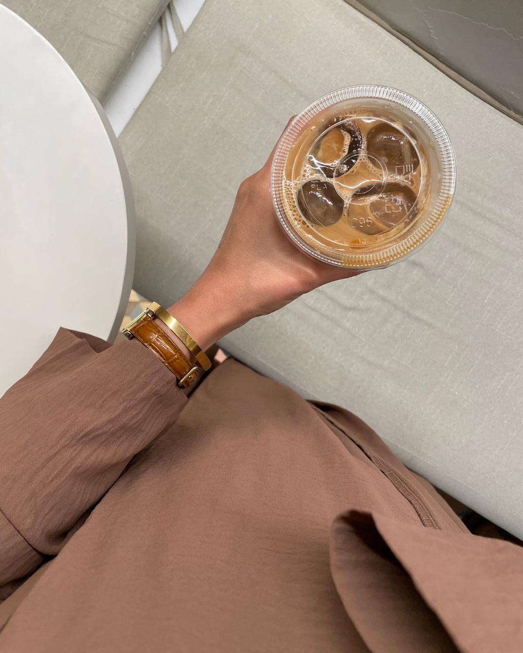 A person in a brown blazer holding a clear plastic cup of iced coffee with ice cubes, wearing a gold-toned bracelet, seated on a light gray upholstered chair.