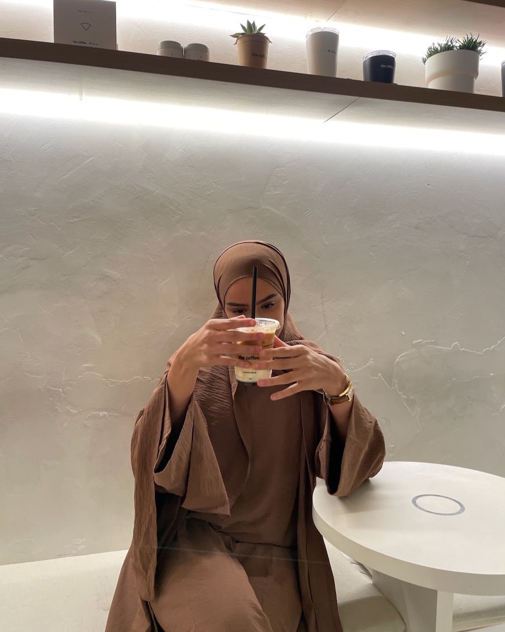 A woman wearing a matching brown hijab and long-sleeved abaya sits at a cafe table, holding a drink with a straw. The background features a textured light gray wall with a shelf holding small potted plants and decorative vases.