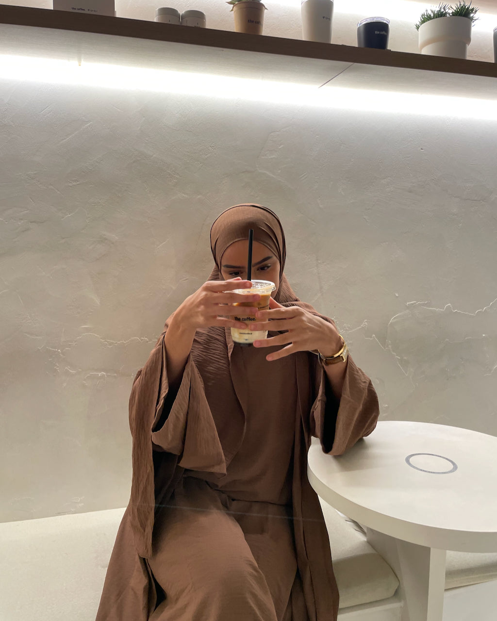 A woman wearing a matching brown abaya and hijab sits in a cafe setting, holding a paper cup in front of her face. The background features a textured light gray wall with a built-in shelf and warm lighting.
