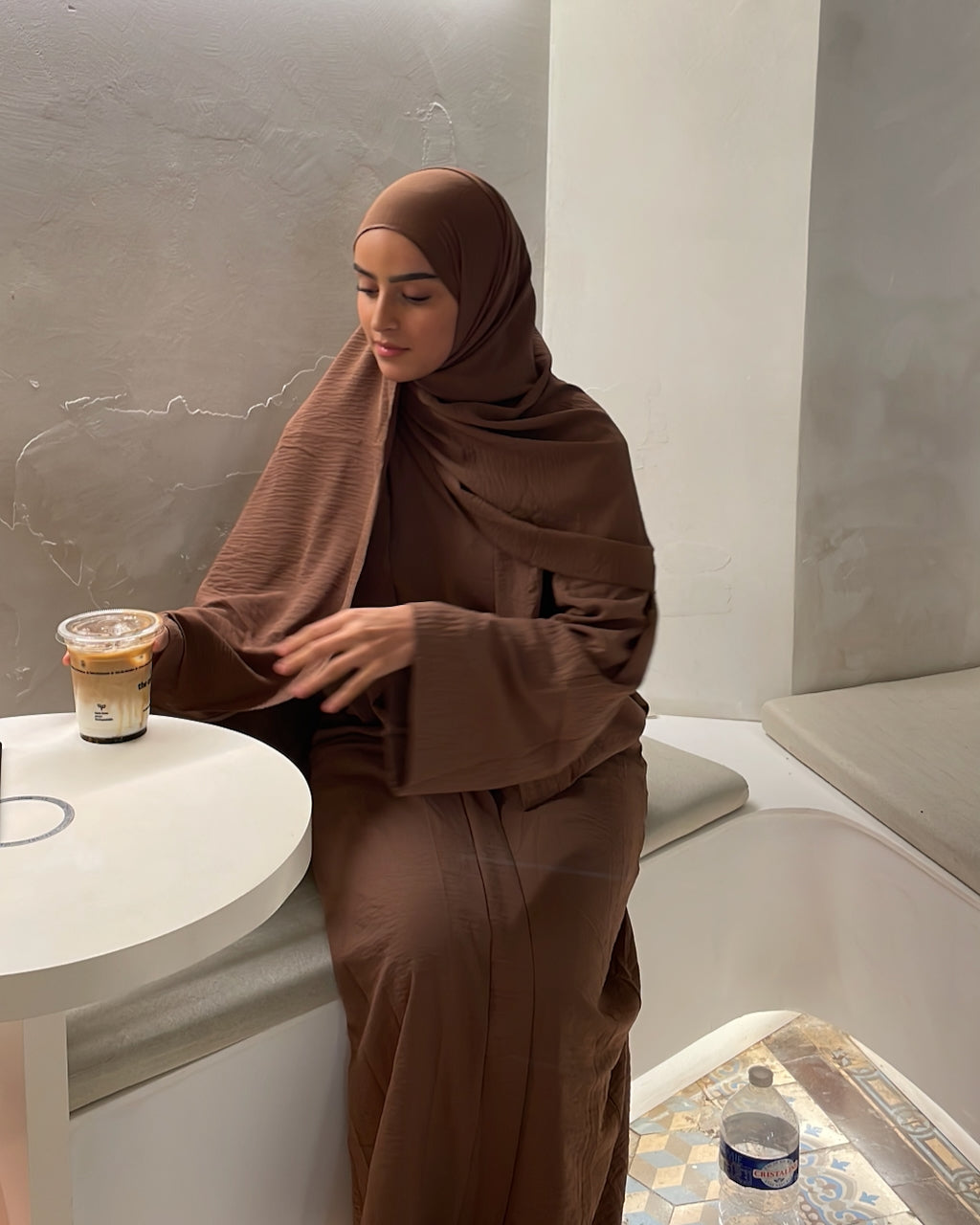 A woman wearing a matching brown hijab and long-sleeve abaya sits at a small white table in a modern indoor setting with a textured gray wall.