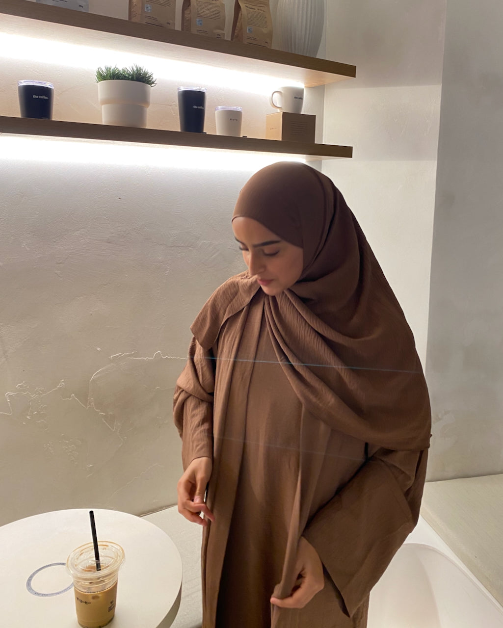A woman wearing a brown hijab and matching long-sleeve abaya stands indoors near a table with an iced coffee. The setting includes a textured wall, wooden shelves with cups and plants, and a white tub.