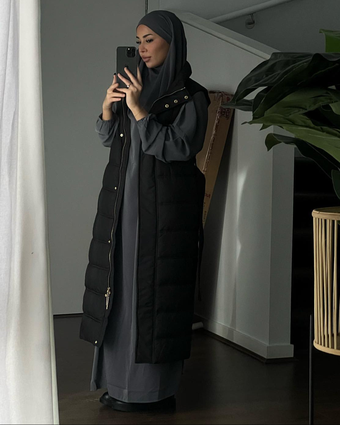 A woman wearing a black puffer vest over a long-sleeved gray dress and a dark gray hijab takes a mirror selfie in a modern room with a large green plant in the foreground.