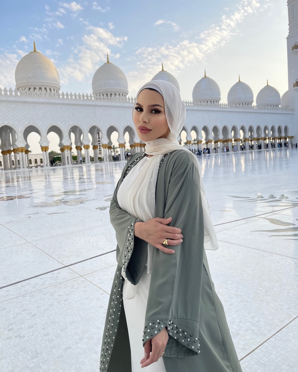 A woman wearing a white hijab and a sage green abaya with white polka dots stands in front of the grand white domes of a mosque under a bright sky.