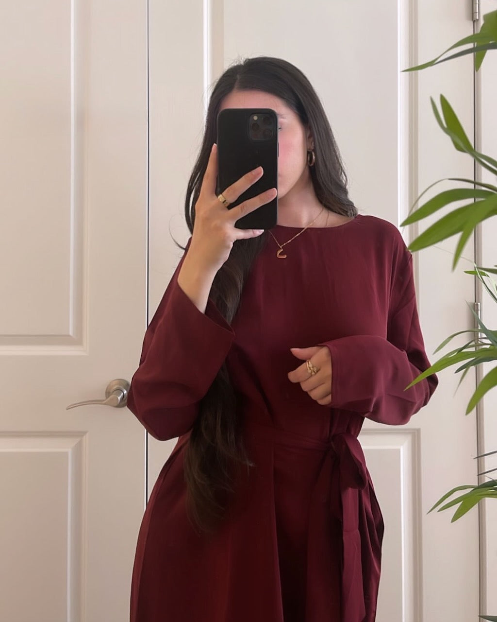 A woman takes a mirror selfie wearing a long-sleeve wrap-style dress in deep burgundy. The dress features a round neckline, long sleeves, and a matching fabric tie belt at the waist.