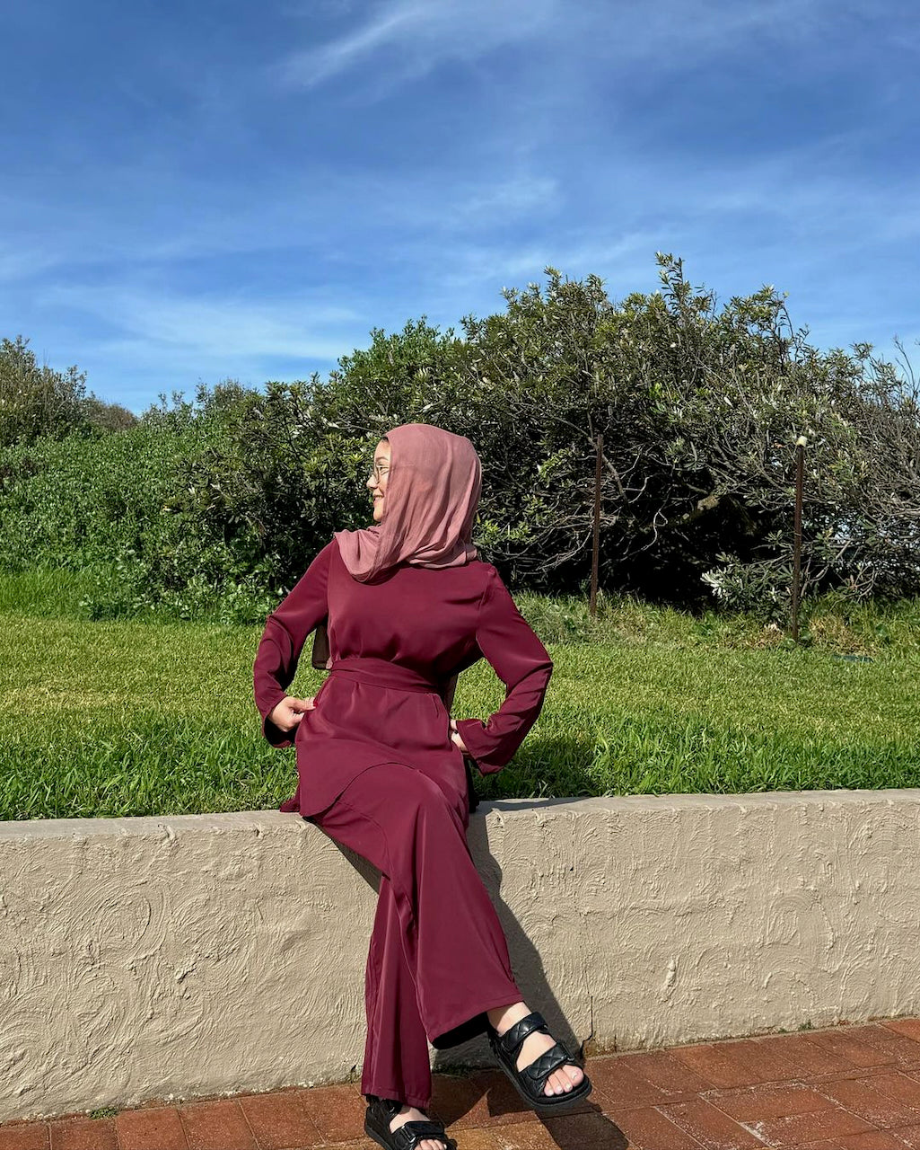 A woman wearing a long-sleeve maroon maxi dress and a dusty pink hijab sits on a low concrete wall outdoors, with green grass, trees, and a blue sky in the background.