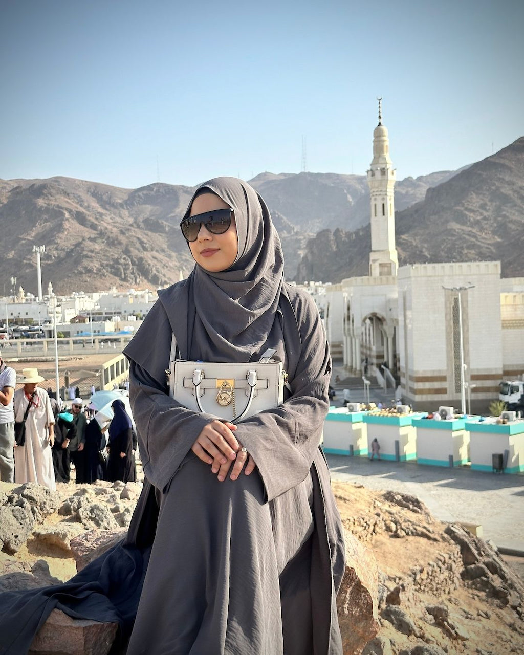 A woman wearing a dark gray hijab and long abaya stands outdoors in front of a white mosque and mountains, holding a light gray structured handbag. Hajj Umrah Abaya Fajr Noor