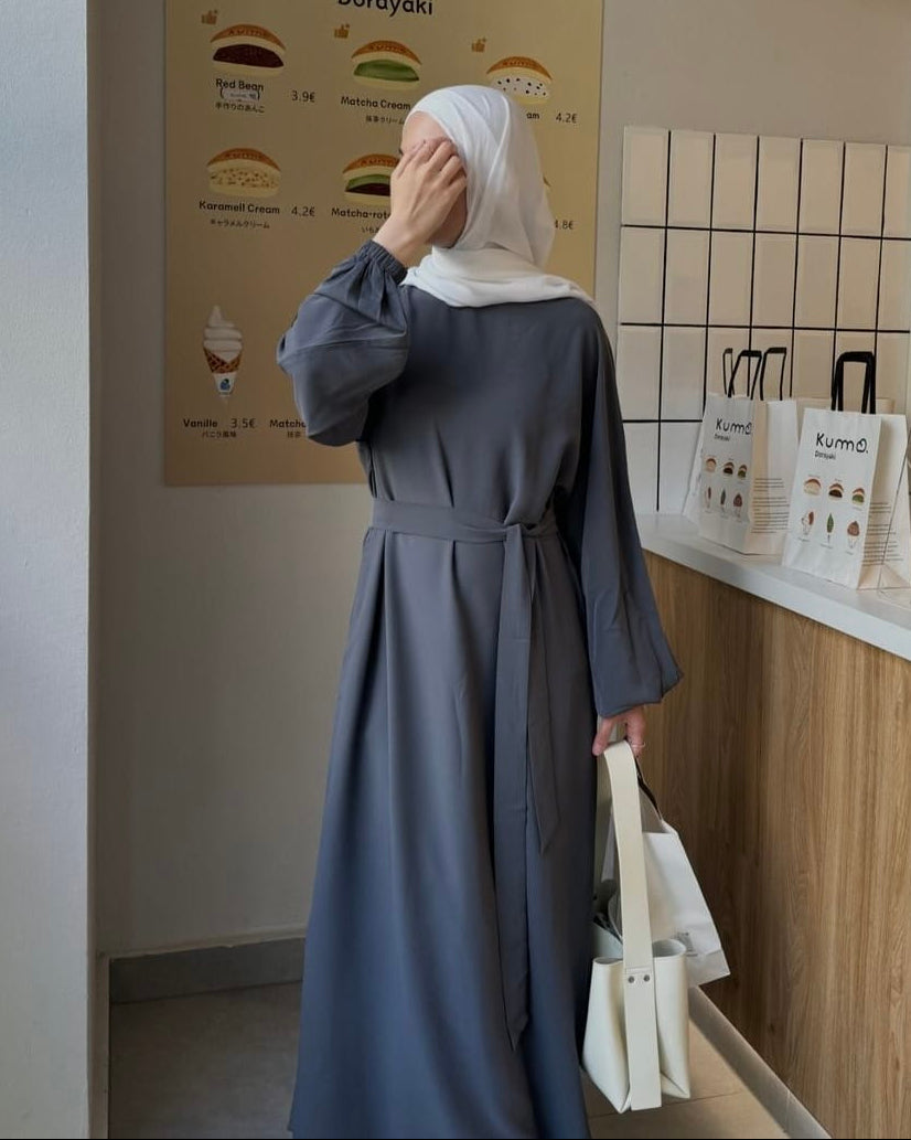 A woman wearing a long, loose-fitting slate blue-gray abaya dress with a matching waist tie and a white hijab stands in a cafe, holding a white tote bag. A Kumo Dorayoki menu poster is visible in the background.