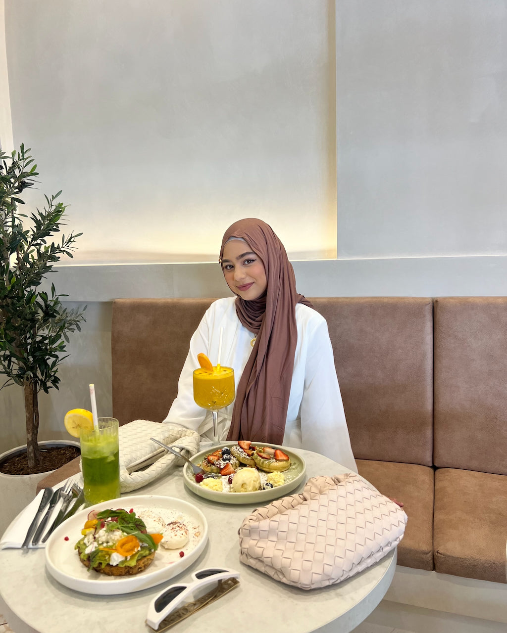 A woman wearing a brown hijab and white long-sleeve top sits at a round table in a cafe, surrounded by food and drinks. She is smiling and holding a yellow drink, with plates of breakfast and a pink woven clutch on the table.