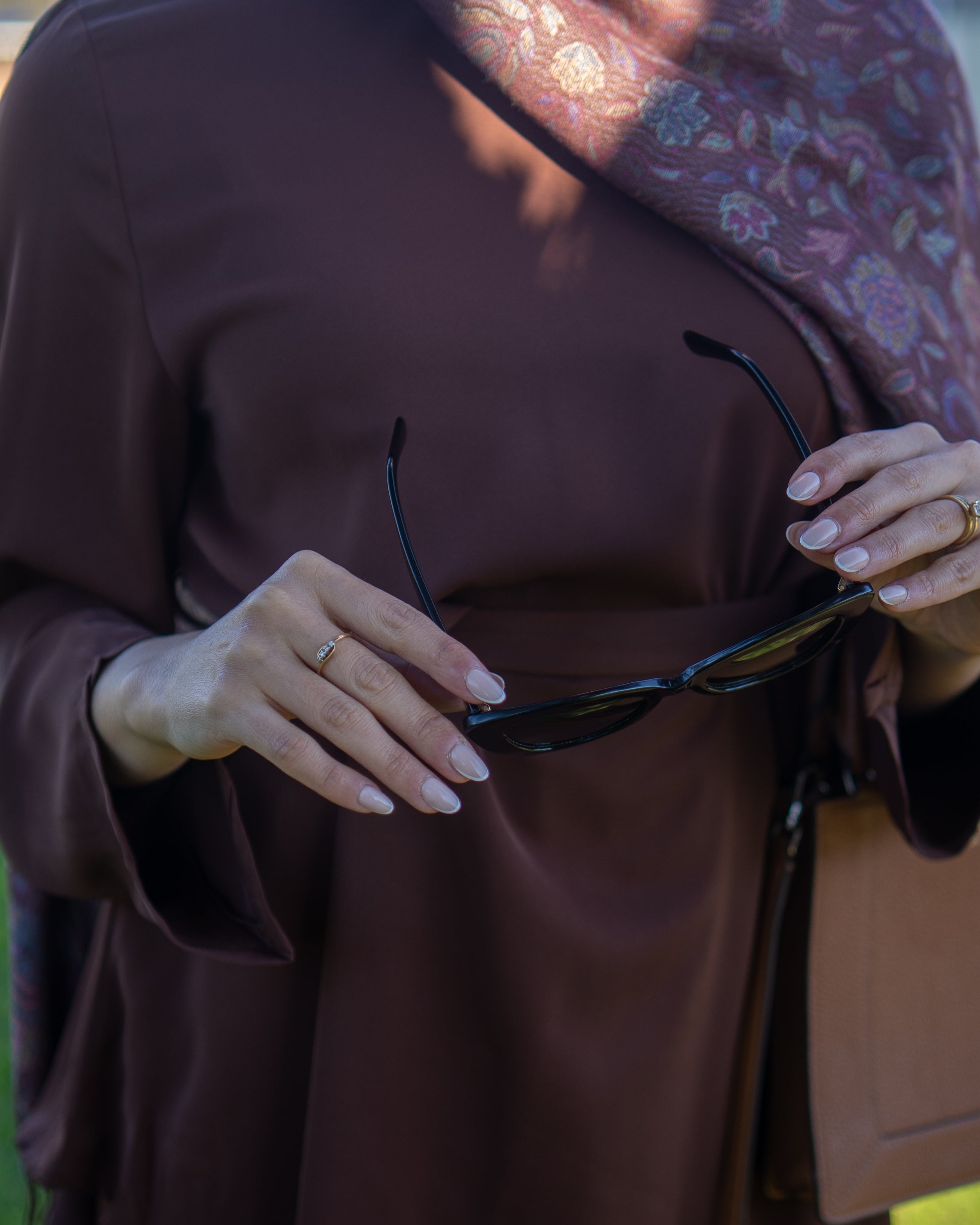 Person wearing a brown garment holding sunglasses with a blurred background