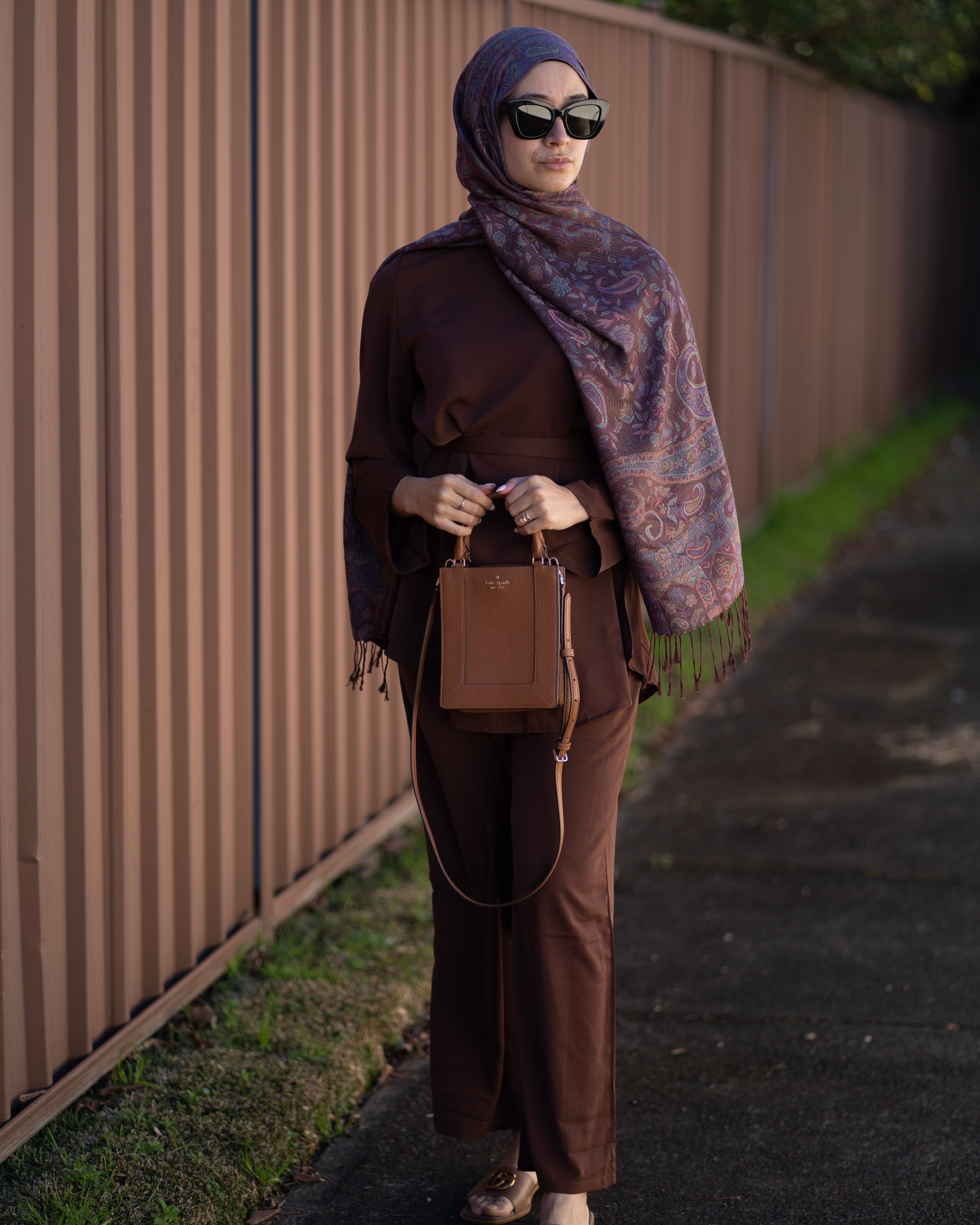 Woman in brown outfit with scarf and sunglasses standing against a wooden fence.