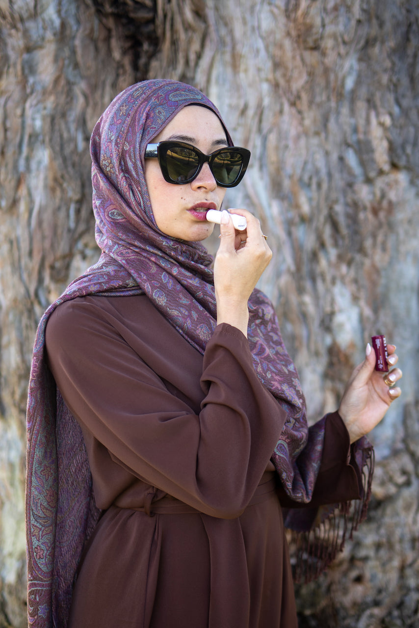 A woman wearing a brown long-sleeve dress and a purple paisley hijab applies lipstick outdoors in front of a textured tree trunk.