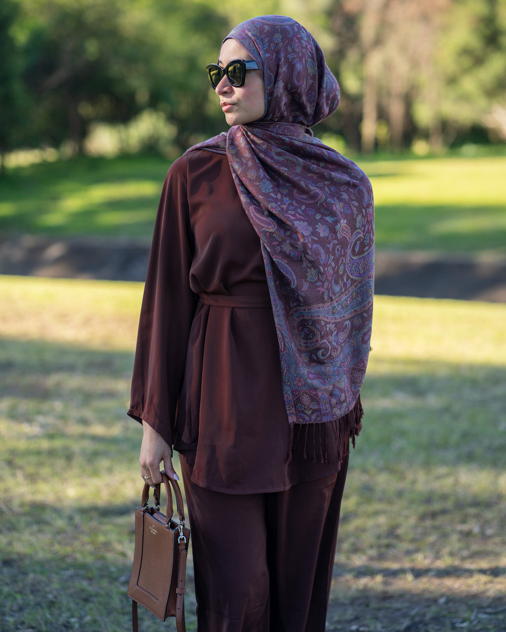A woman wearing a dark brown long-sleeve tunic and matching pants, styled with a purple paisley-patterned hijab and sunglasses, holding a brown handbag in an outdoor park setting.