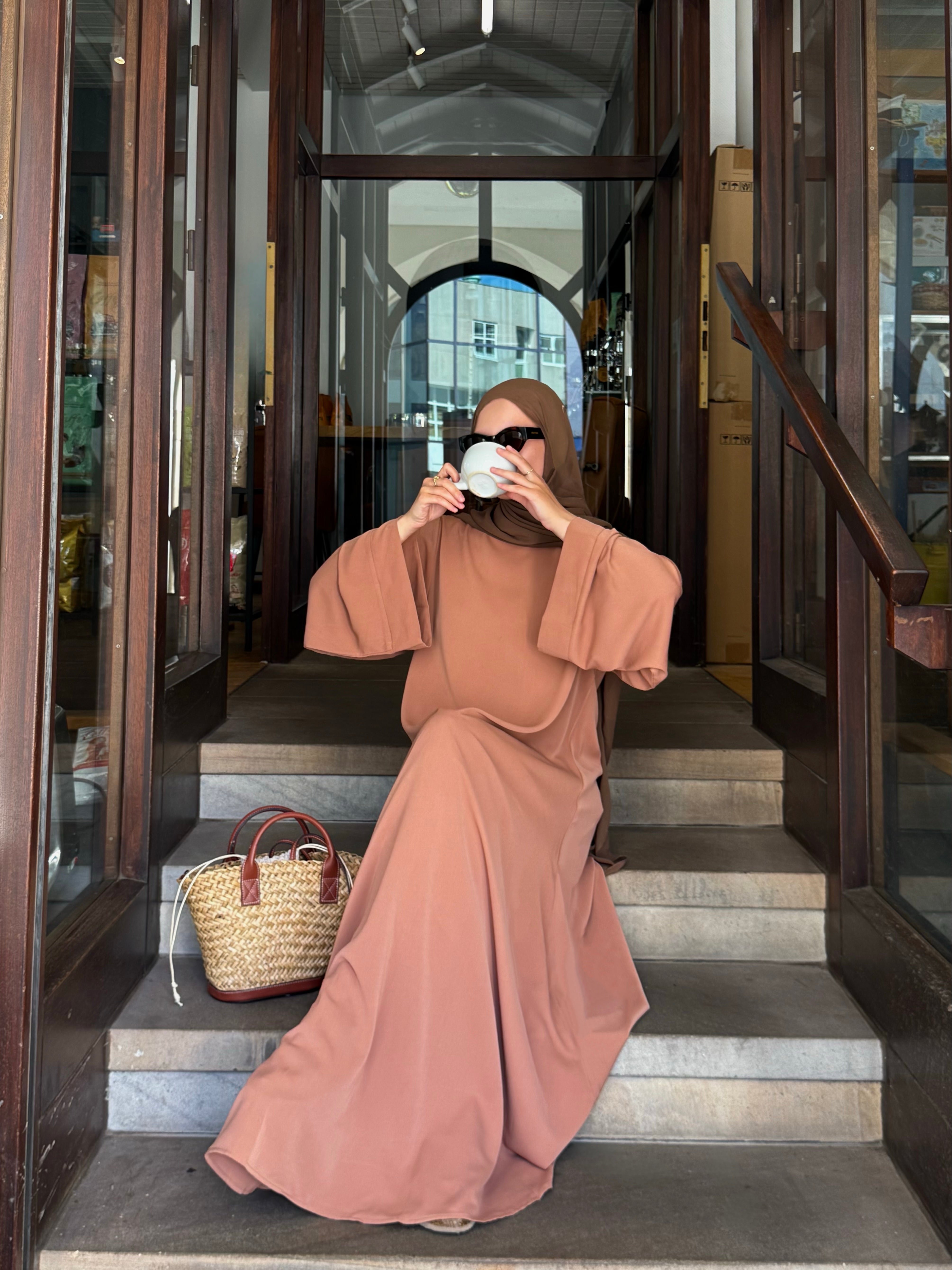 Woman in a long, light brown dress sitting on steps inside a building.