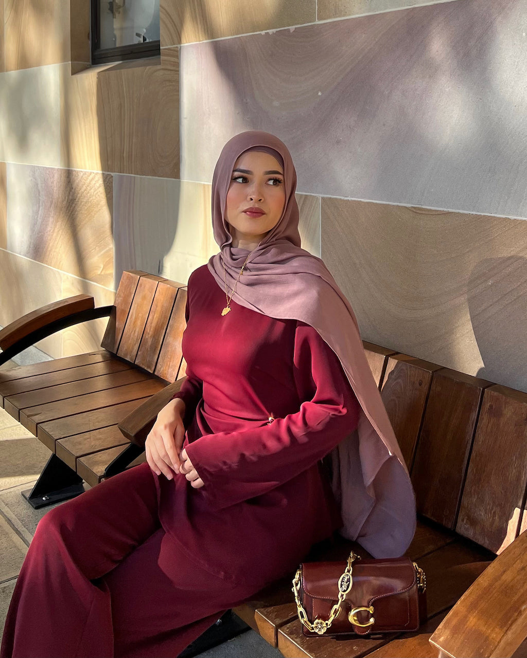 A woman wearing a matching deep burgundy long-sleeve dress and dusty mauve hijab sits on a wooden bench. She is accessorized with a small gold necklace and a brown leather shoulder bag with a gold chain, set against a background of warm-toned stone tiles.