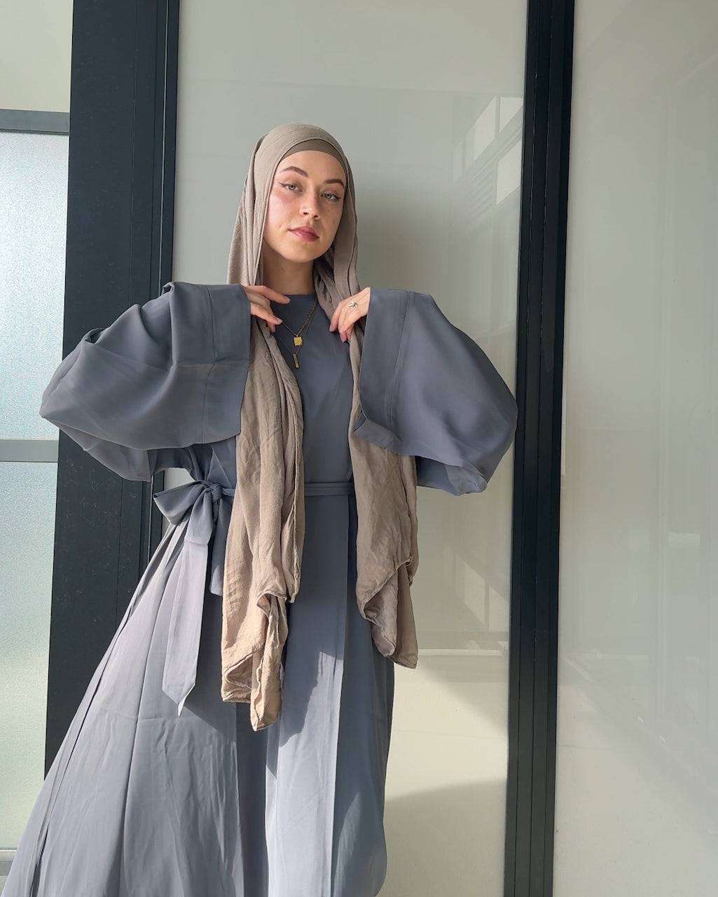 A woman wearing a slate blue-gray modest outfit with a matching hijab, featuring a long-sleeved open-front outer layer with wide sleeves and a draped scarf, standing in front of a glass door.