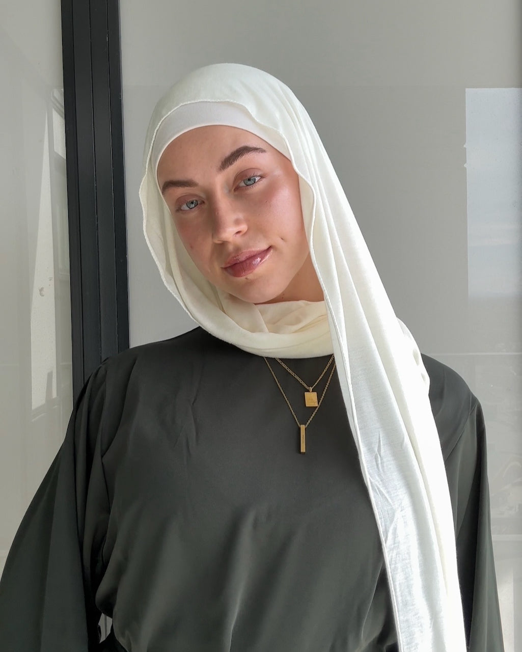A woman wearing a white hijab and a dark gray abaya, accessorized with gold necklaces, standing indoors.