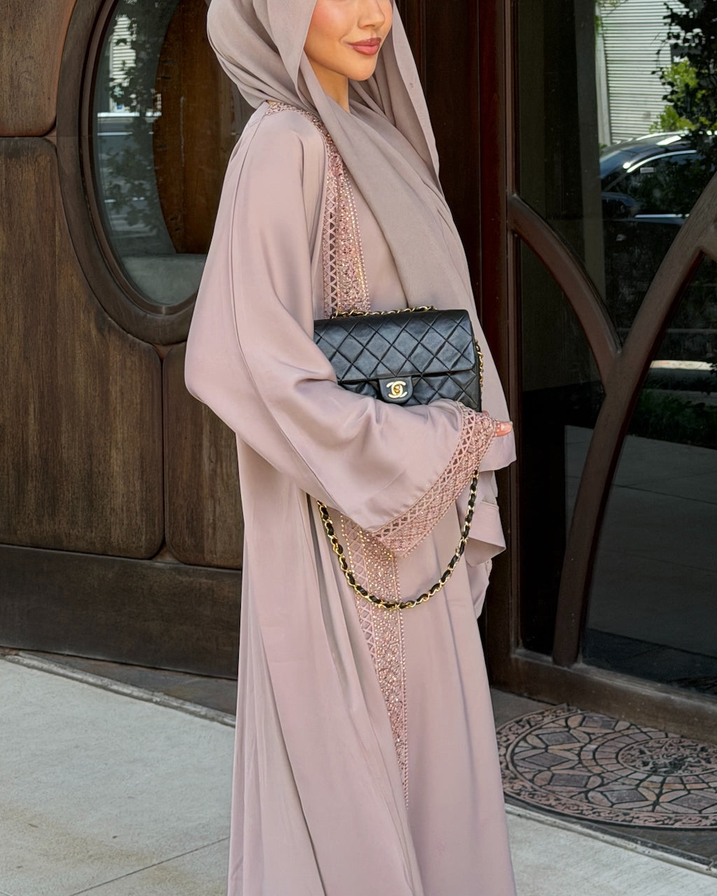 A woman wearing a flowing dusty pink abaya and matching hijab, holding a black quilted Chanel-style handbag with a gold clasp and chain strap, standing in front of a wooden building entrance. - Fajr Noor