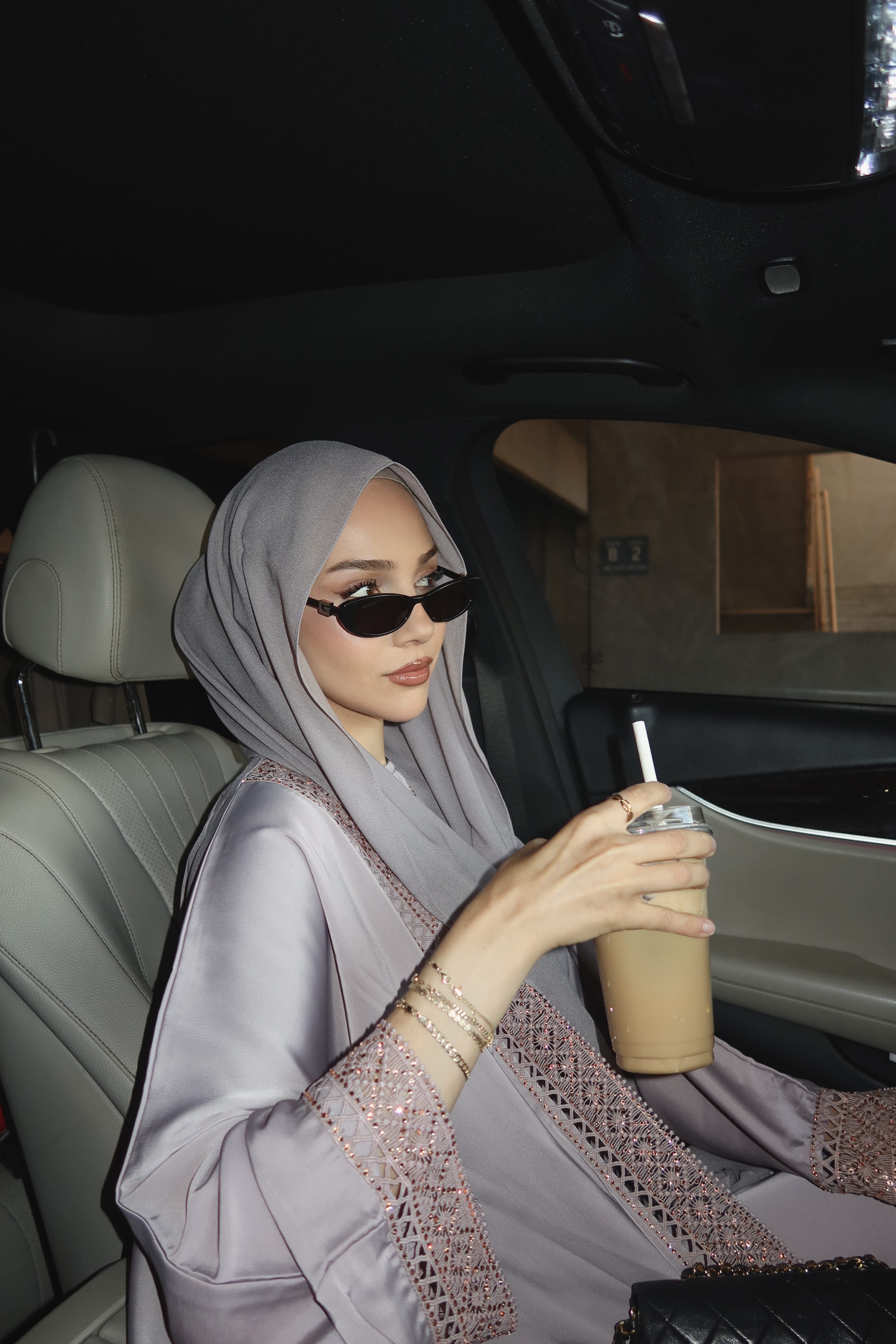 A woman wearing a light gray hijab and a matching gray embroidered abaya sits inside a car, holding an iced coffee.