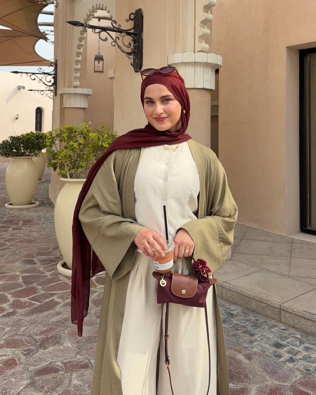 A woman wearing a beige long-sleeve kaftan over a moss linen abaya, styled with a deep burgundy hijab and a matching small crossbody bag, standing outdoors in front of a beige building.