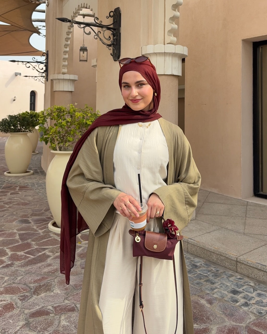 A woman wearing a beige long-sleeve abaya and a deep maroon hijab stands outdoors in front of a beige building. She is holding a small maroon crossbody bag and a coffee cup.