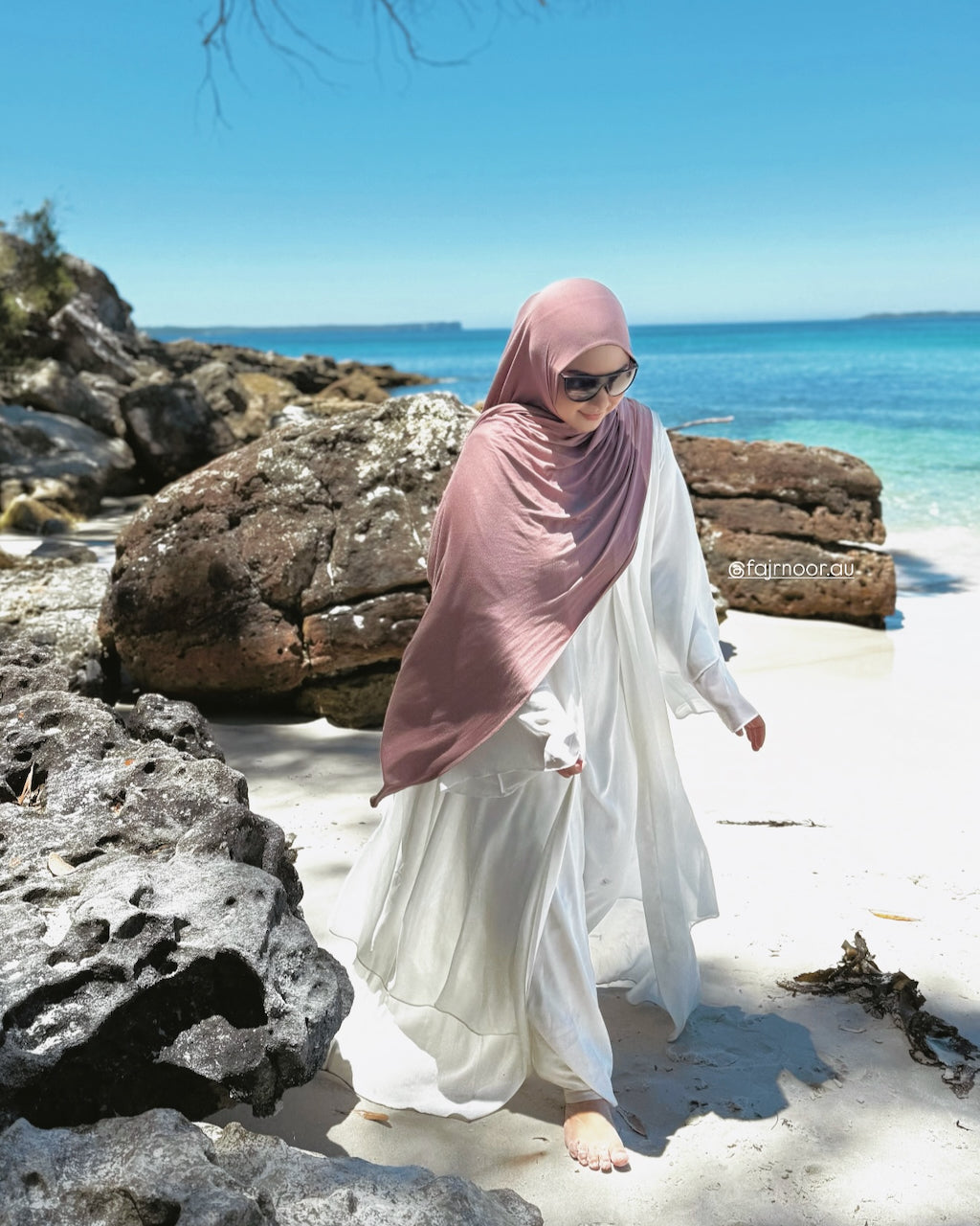 A woman wearing a dusty rose hijab and a long white dress walks barefoot on a sandy beach with large rocks and turquoise water in the background.