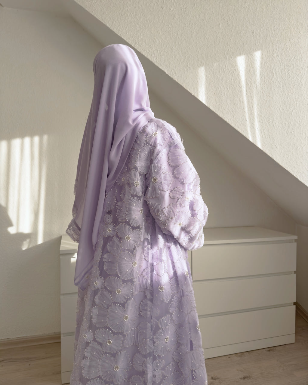 A person wearing a light lavender floral abaya and matching hijab, photographed from behind in a sunlit room with a white dresser and slanted ceiling.