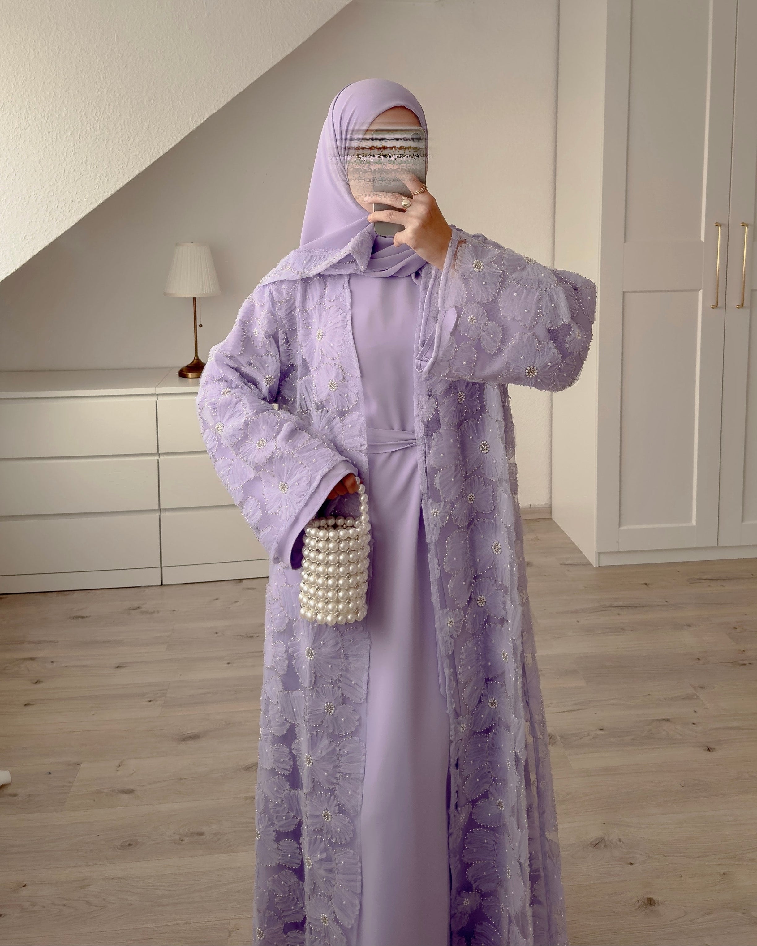 Person wearing a long light purple dress with a hood in a room with white walls and wooden floor.