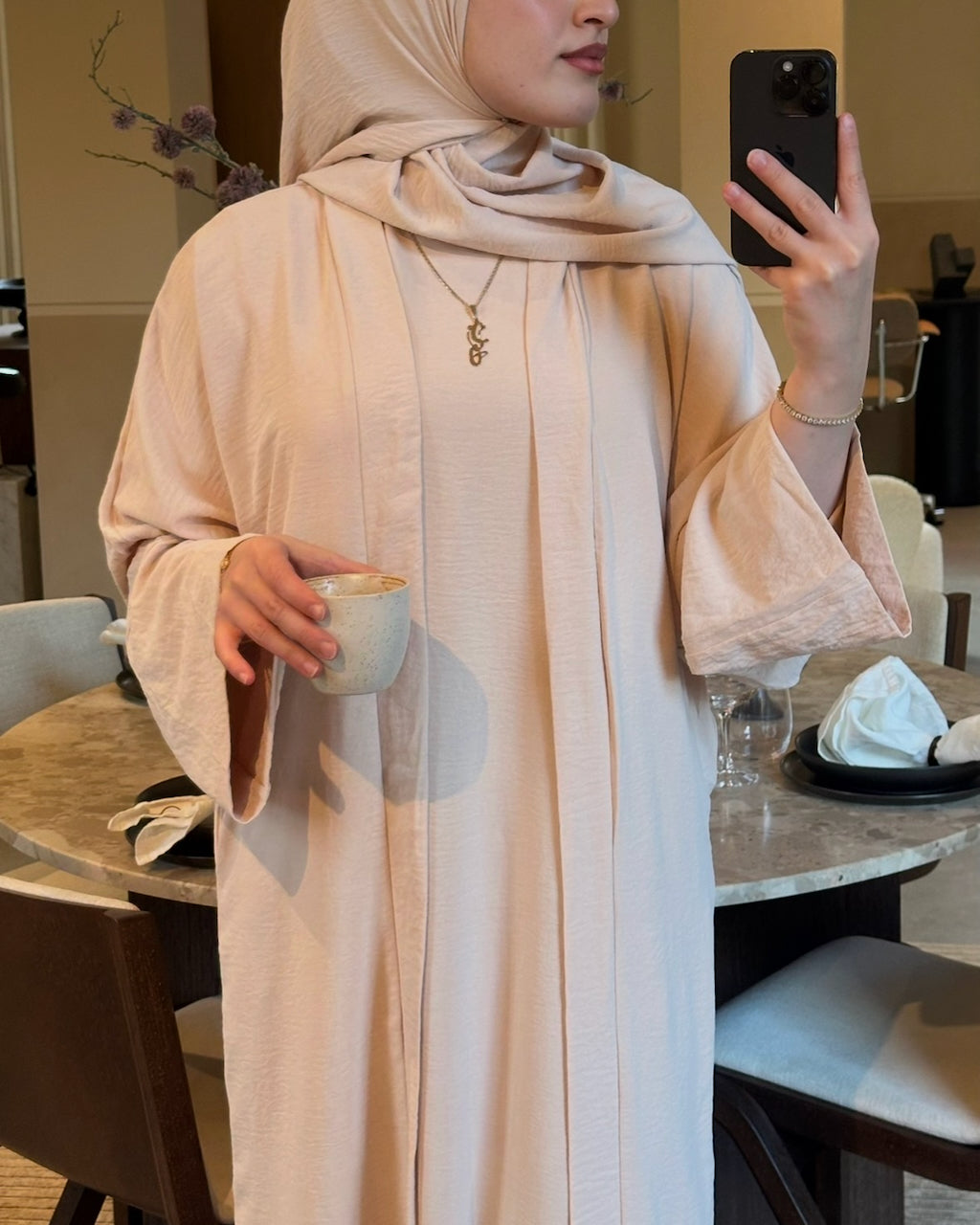 A woman wearing a light beige textured abaya and matching hijab takes a mirror selfie in a dining area, holding a small ceramic cup.