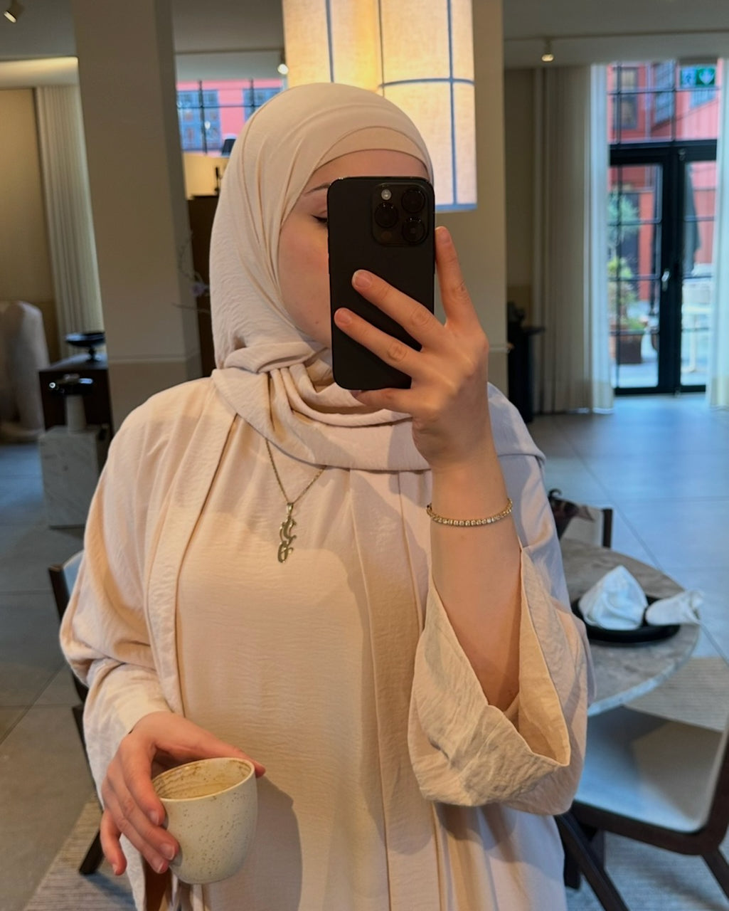 A woman wearing a light beige hijab and matching long-sleeve blouse takes a mirror selfie in a modern indoor setting, holding a small cup.