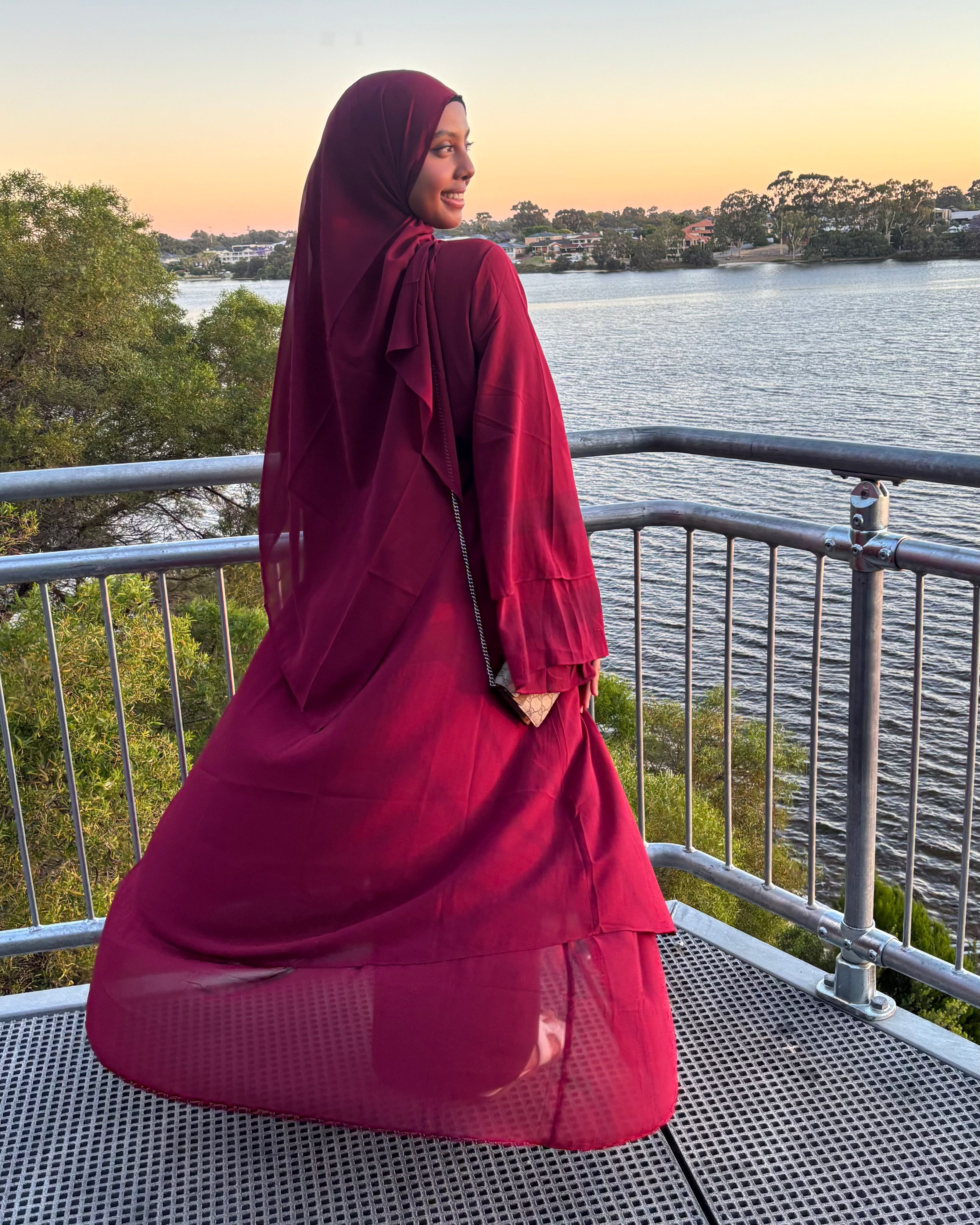 Woman in a red hijab and dress standing on a balcony overlooking a lake at sunset.