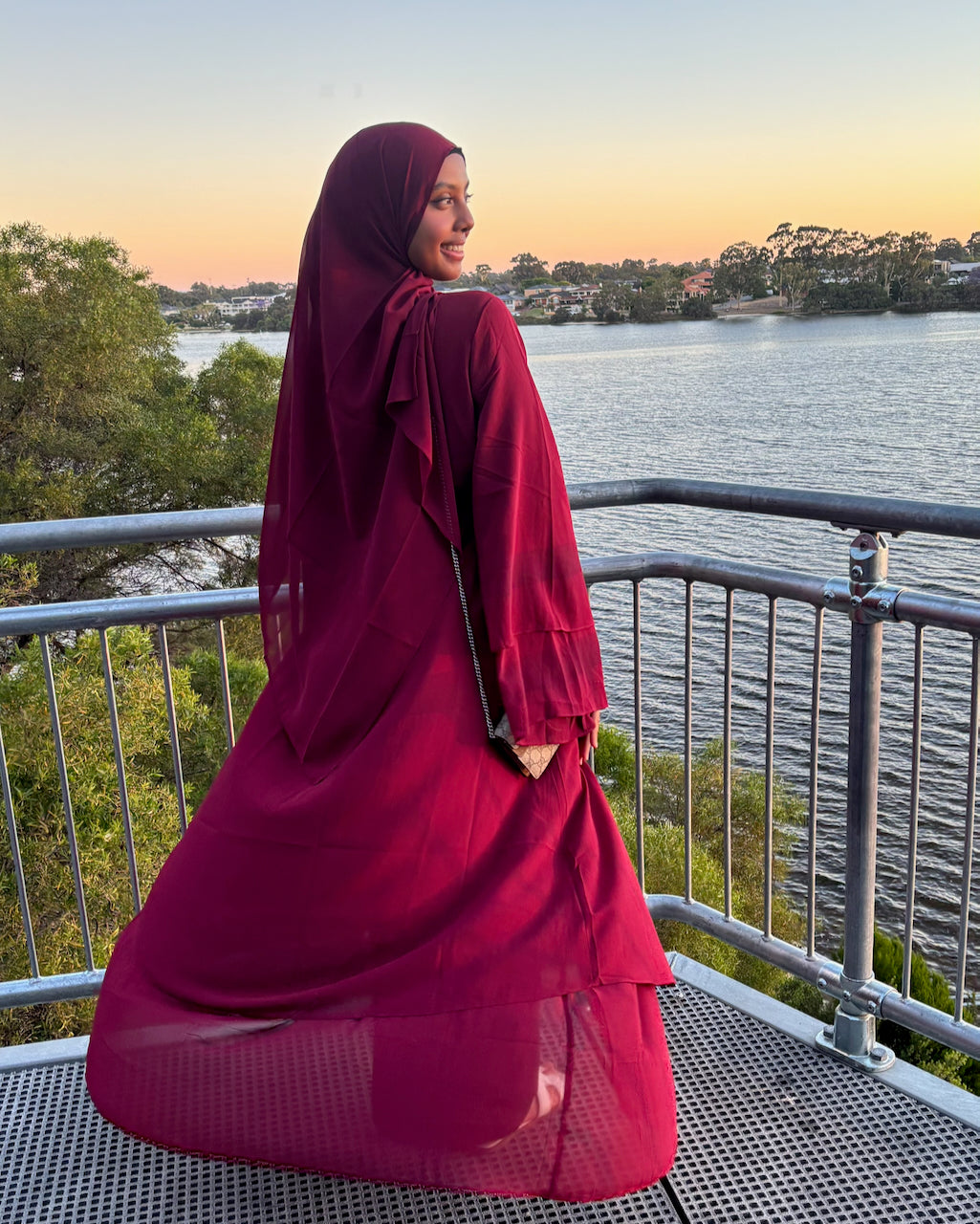 A woman wearing a deep burgundy hijab and matching long-sleeve abaya stands on a metal walkway overlooking a lake at sunset.