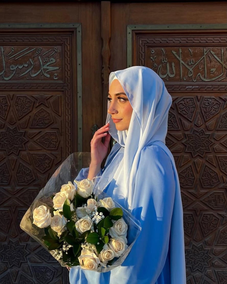 A woman wearing a light blue hijab and matching long-sleeve dress stands in front of an ornate dark brown wooden door with Arabic calligraphy, holding a bouquet of white roses.