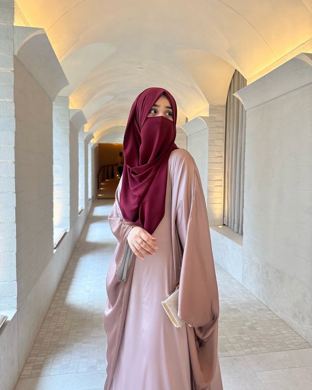 Woman in a pink abaya and maroon hijab standing in a sunlit hallway with arches.