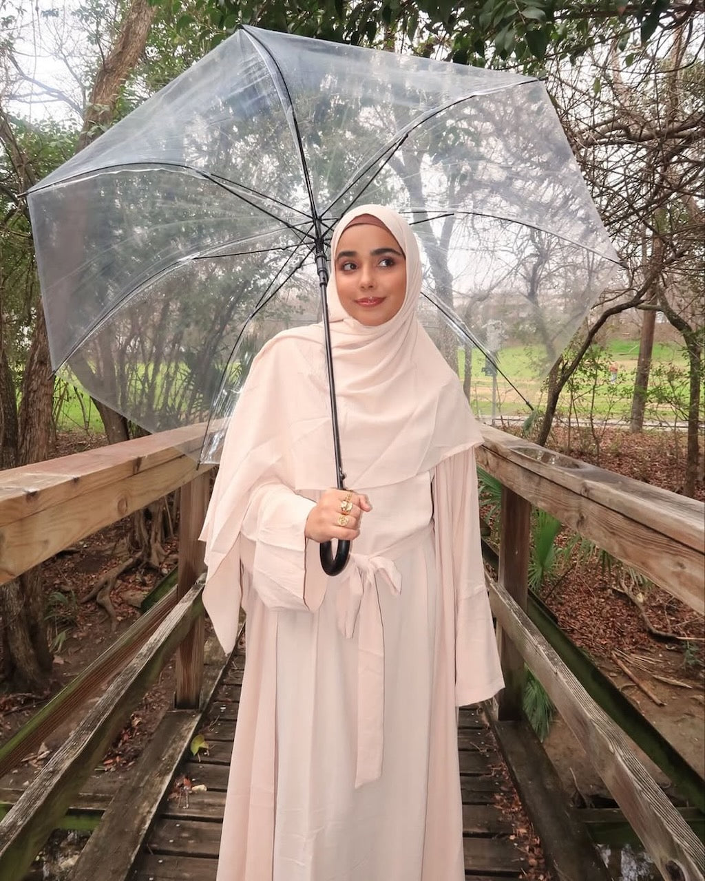 A woman wearing a light blush pink abaya and matching hijab stands on a wooden boardwalk holding a clear transparent umbrella. The background shows a wooded outdoor setting with trees and a path.