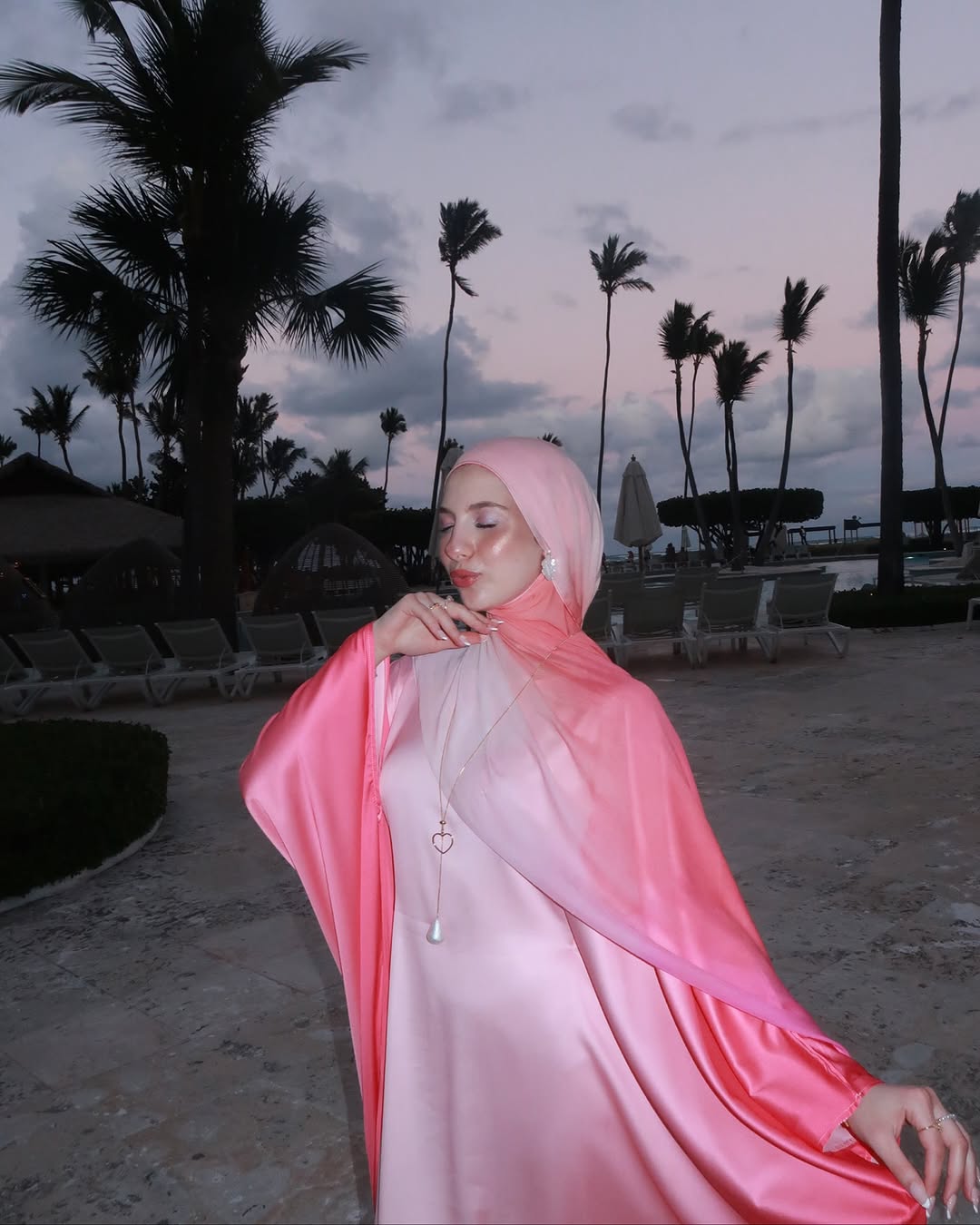 Woman in a pink hijab and dress standing on a beach with palm trees in the background.