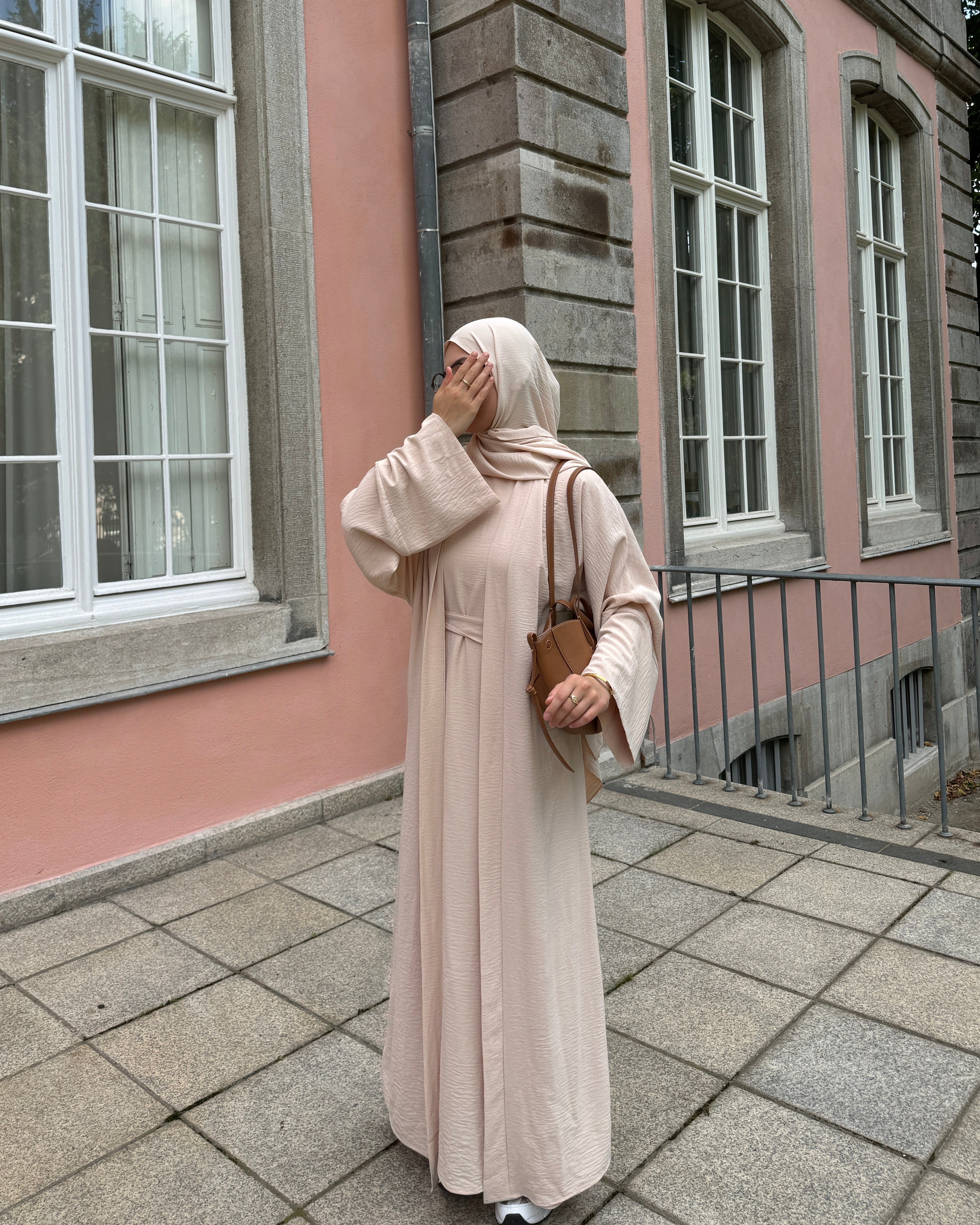 Woman in a long beige dress standing in front of a pink building with white windows.