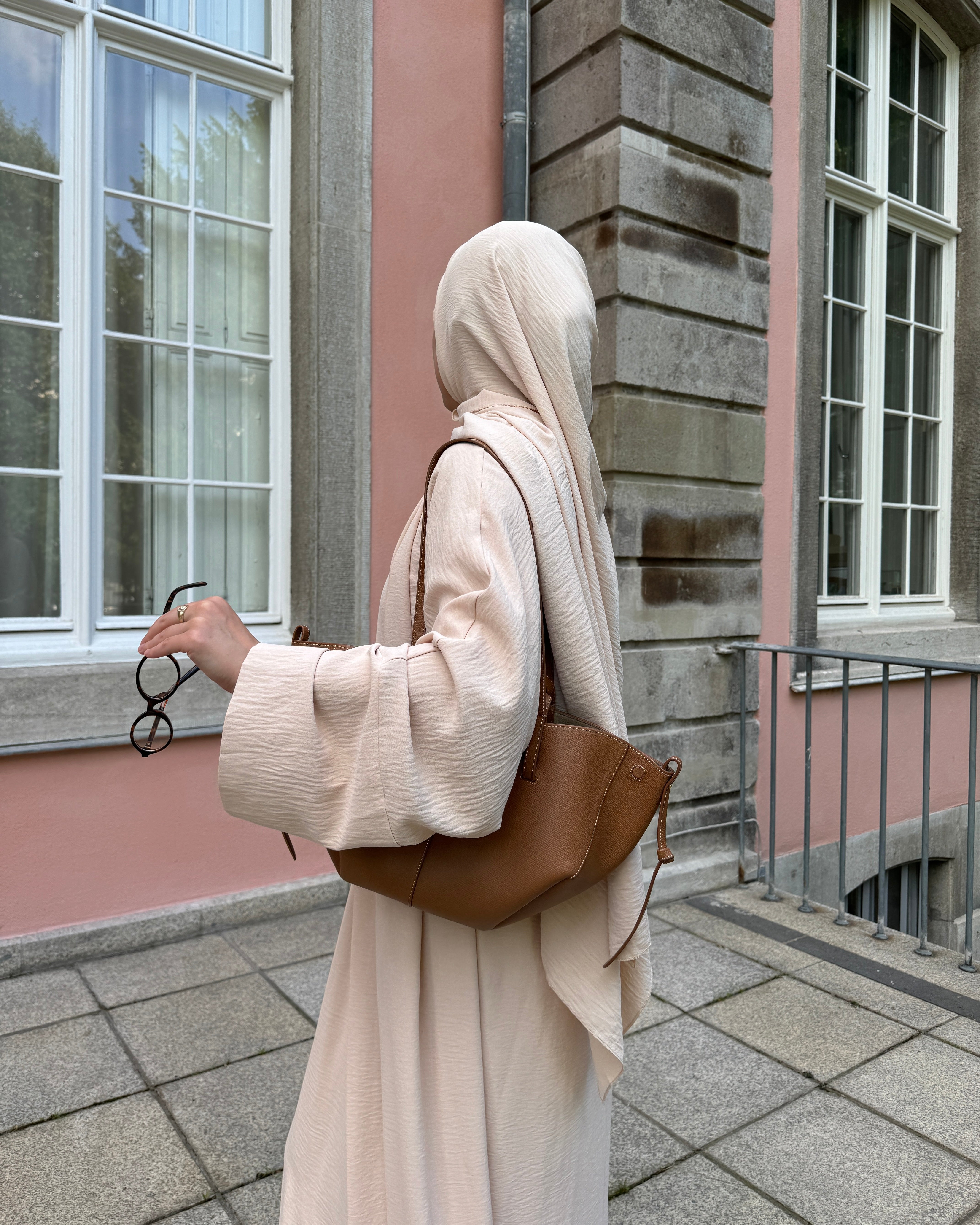 Person wearing a beige hijab and holding a brown bag in front of a building.