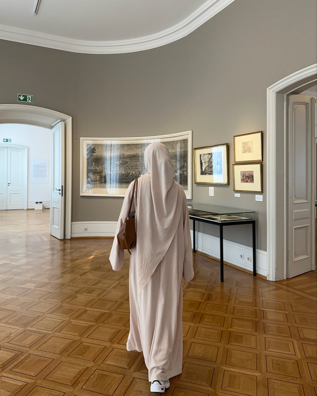 A person wearing a light beige abaya and matching head covering walks through a gallery with wood parquet floors and framed art on gray walls.