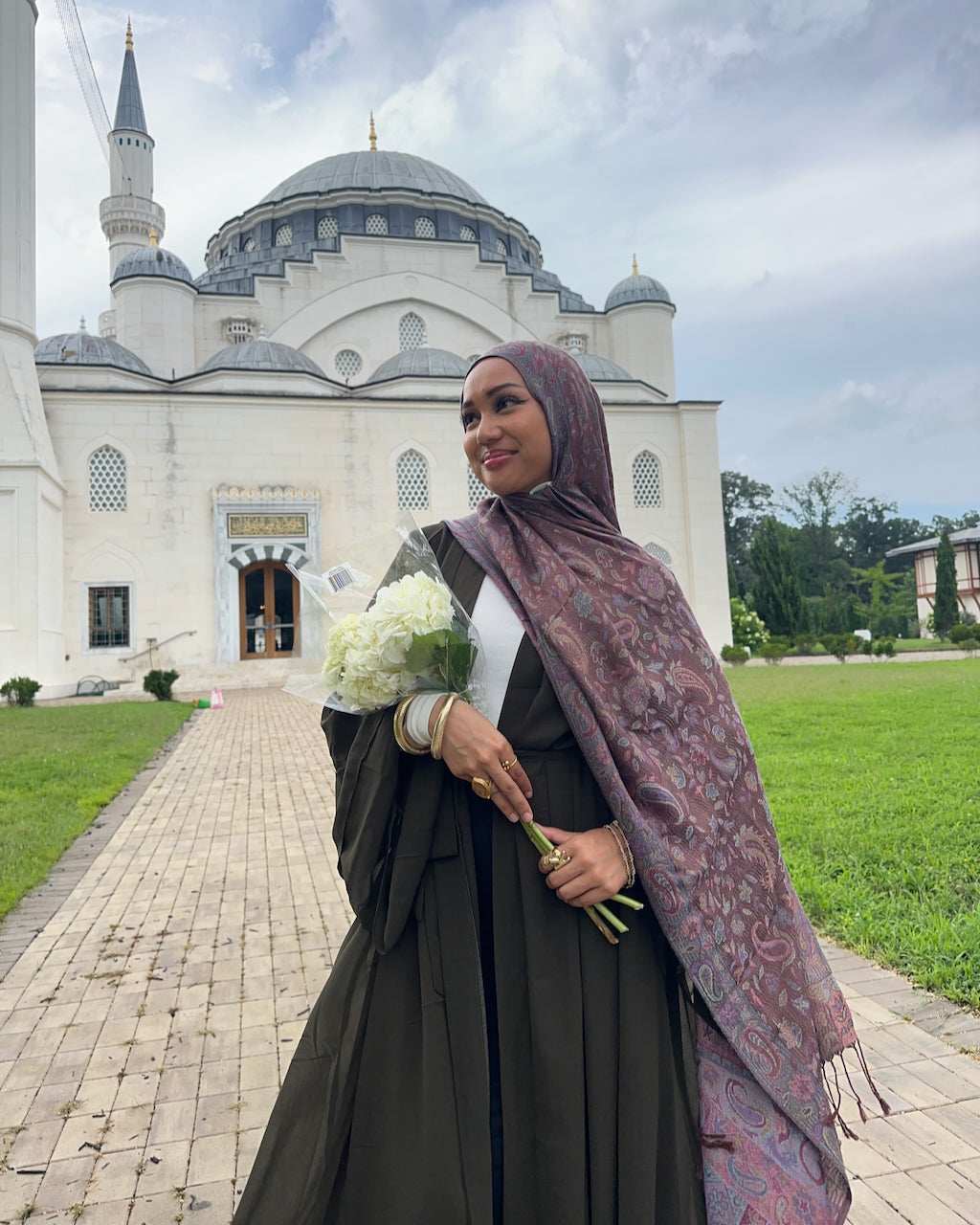 A woman wearing a dark brown abaya and a mauve paisley hijab stands outdoors in front of a large white mosque with minarets, holding a bouquet of white flowers.