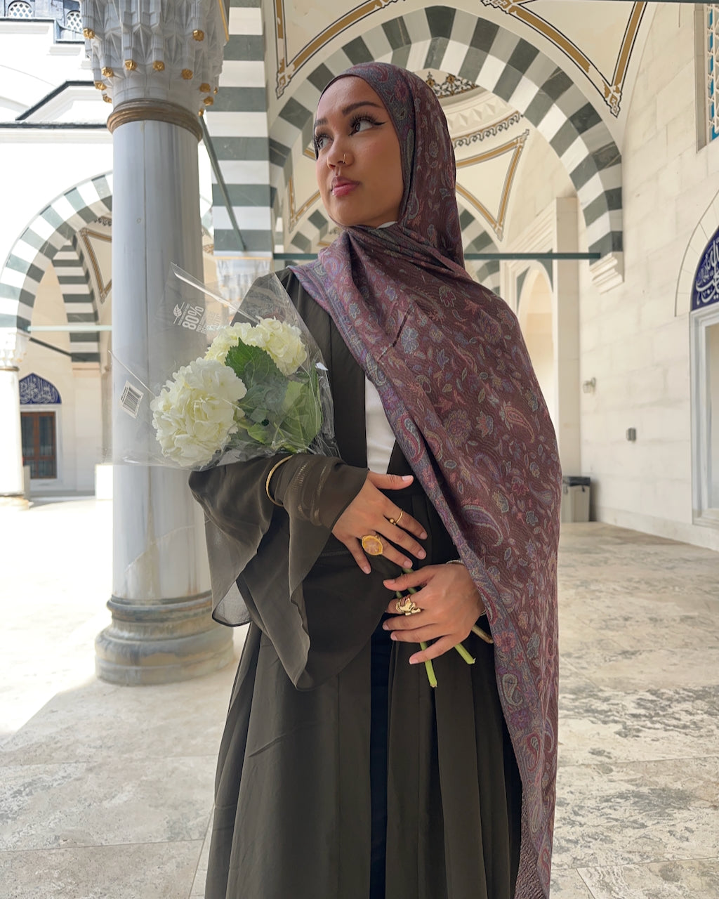A woman wearing a brown and purple paisley-patterned hijab and a dark olive green dress stands in a historic stone courtyard with arches, holding a bouquet of white flowers.