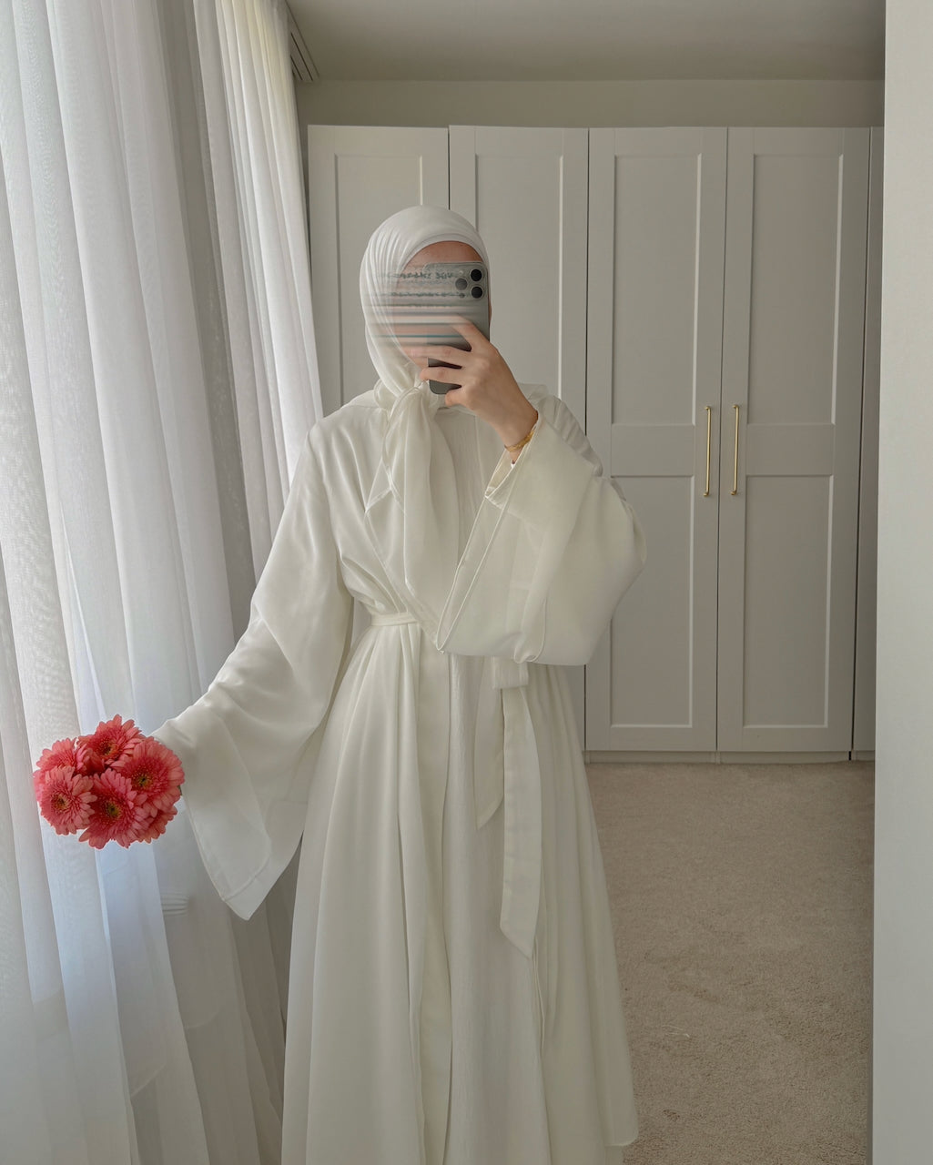 A person takes a mirror selfie wearing a long white robe with wide flowing sleeves and a matching head covering, standing indoors in front of white closet doors and sheer curtains, while holding a small bouquet of pink flowers.