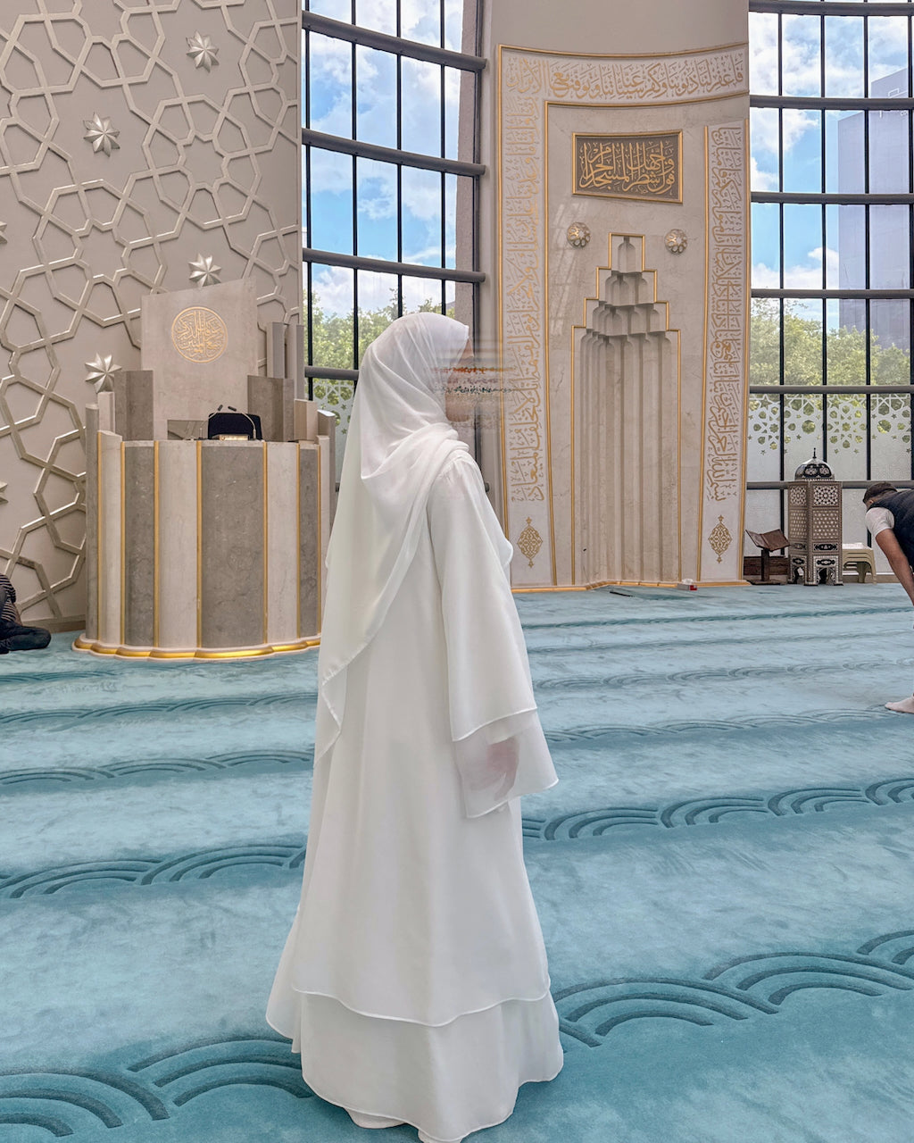 A woman wearing a flowing white modest dress and hijab stands on a blue patterned prayer carpet inside a large, ornate mosque with tall windows and decorative walls.