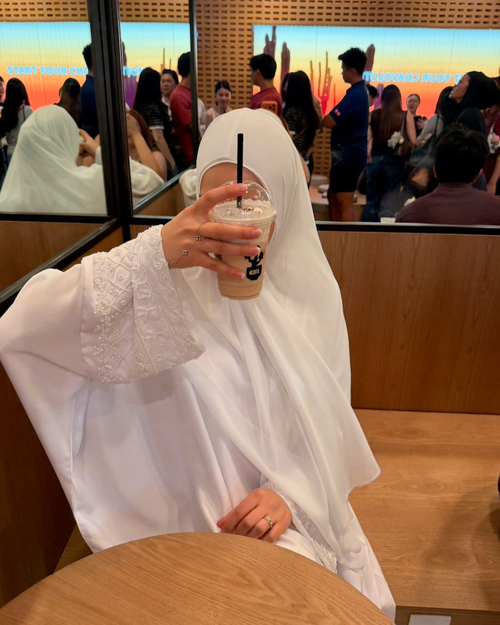 A person wearing a white modest garment and head covering sits in a cafe, holding an iced coffee in front of their face. The background shows other people and cafe seating.