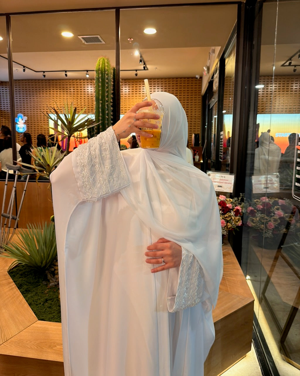 A woman wearing a white modest garment with sheer sleeves and a matching head covering takes a mirror selfie in a modern indoor space with glass walls and plants.