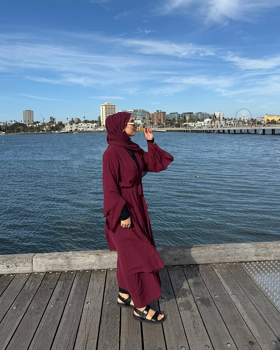 A woman wearing a long burgundy abaya and matching hijab stands on a wooden pier with a body of water and city skyline in the background under a blue sky.